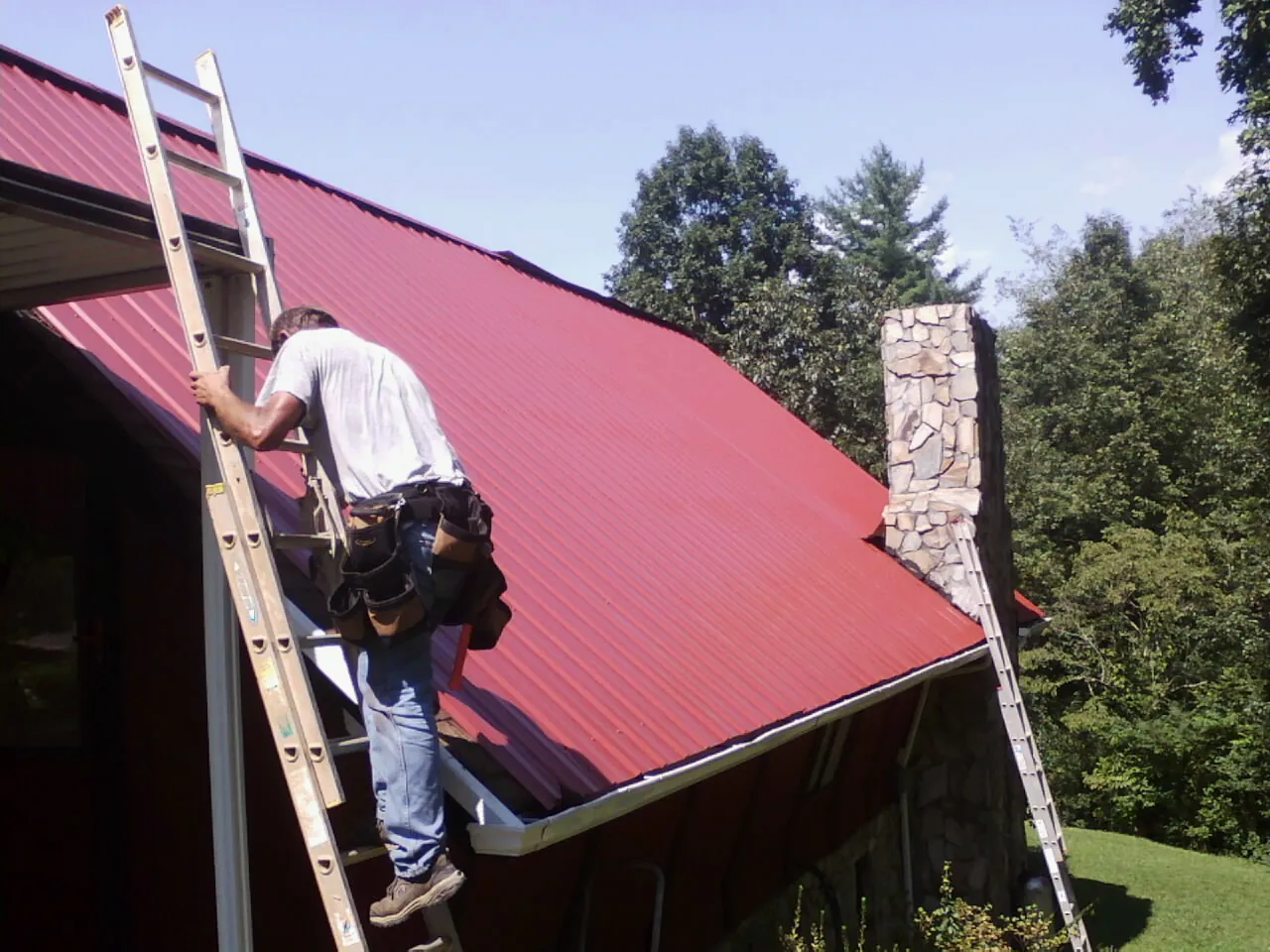 Roofer on a ladder beside a red metal roof and stone chimney — Soffit & Fascia Repair by Roofing Brothers in Sabula